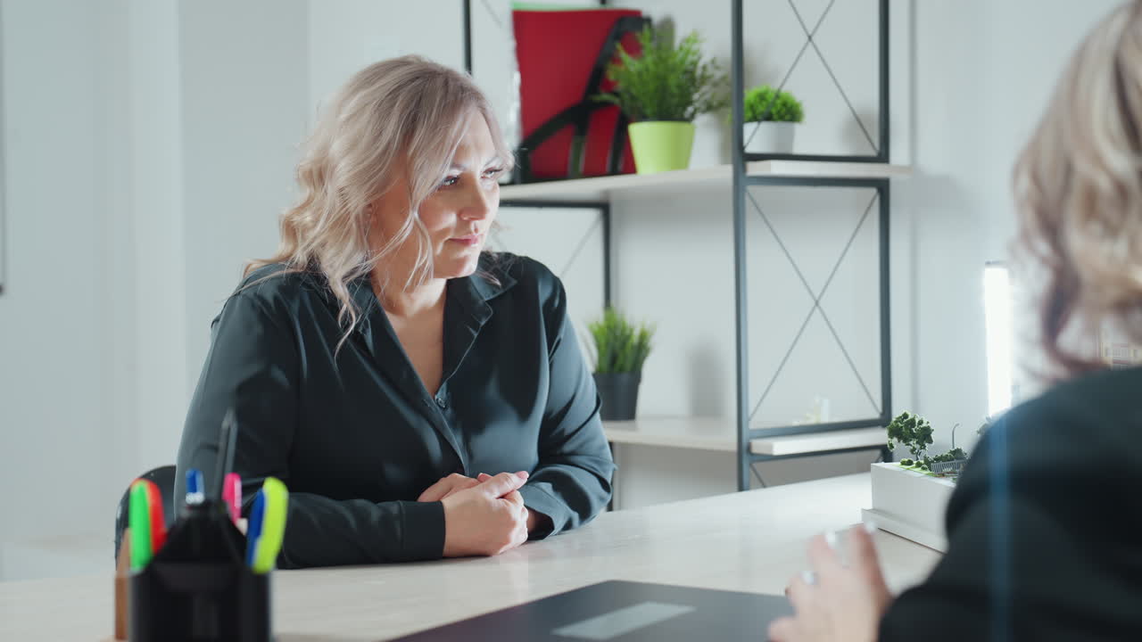 client paying attention to what business expert is telling her with bright light by side seated across office table with hands clasped indoor setting includes shelf with green plants