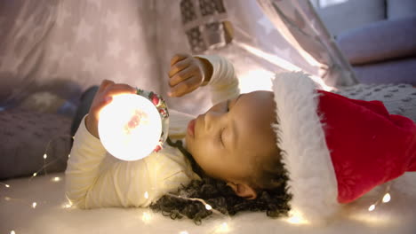 feliz chica afroamericana con sombrero de navidad sosteniendo un globo de nieve brillante en la tienda de la manta, cámara lenta