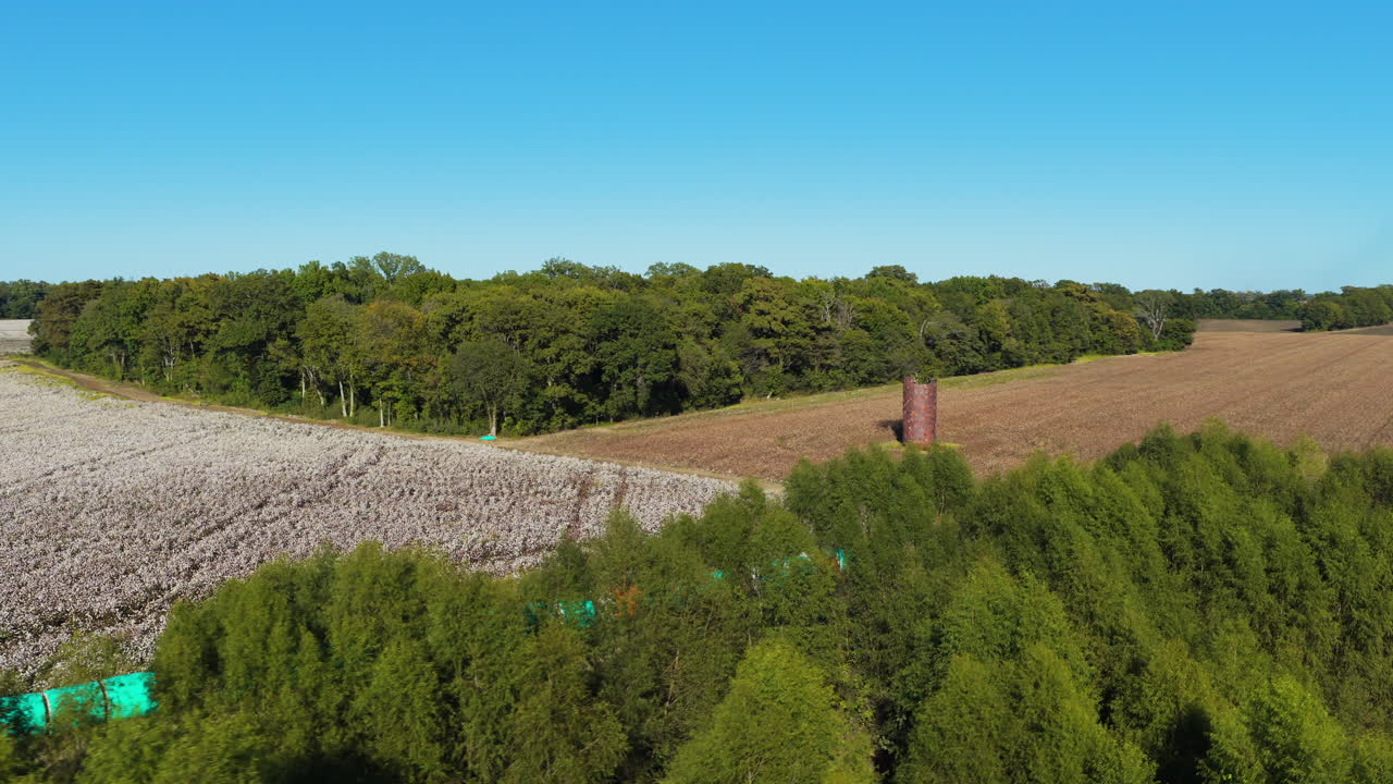 Colorful wrapped cotton bales line a road beside white Mississippi fields and trees, aerial orbit