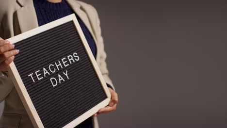 Close-Up-Studio-Shot-Of-Female-Teacher-Standing-Against-Grey-Background-Holding-Notice-Board-Reading-Teachers-Day