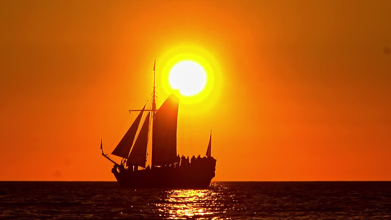 Historic sailing ship glides across glowing orange water under massive sun in slow motion
