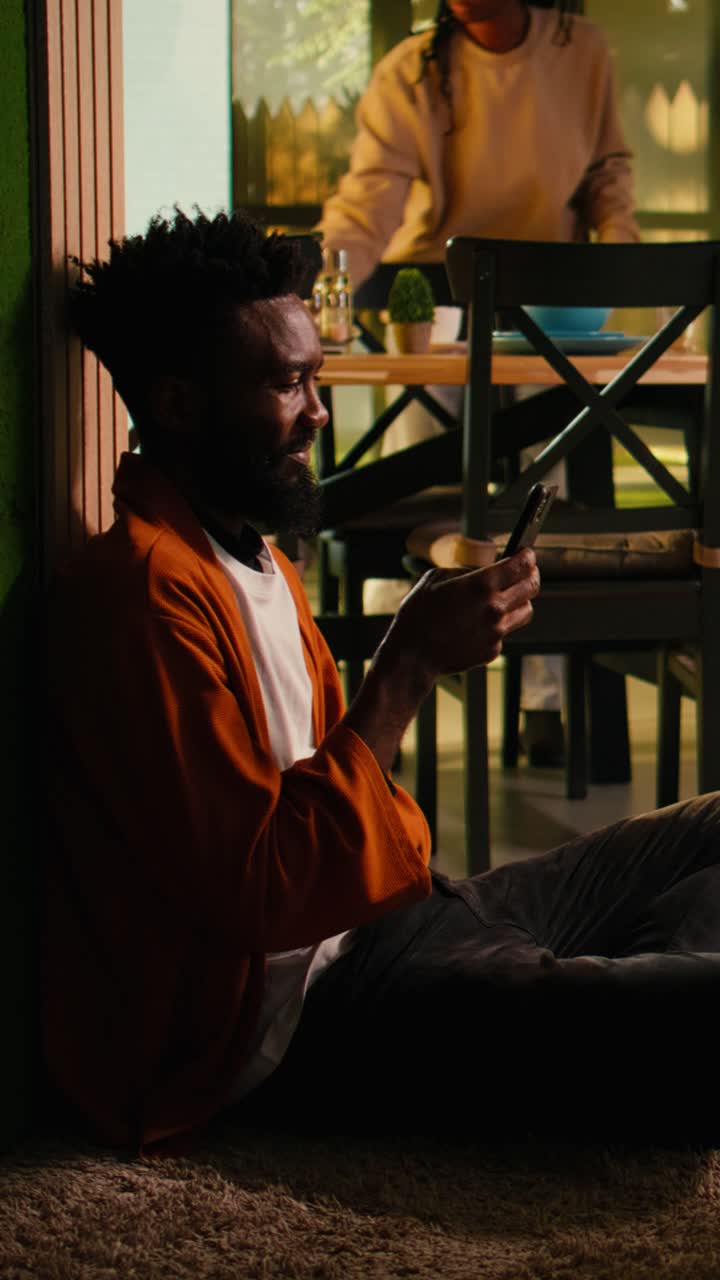 Vertical Video Peaceful indoor moment of black man scrolling on smartphone sitting on floor