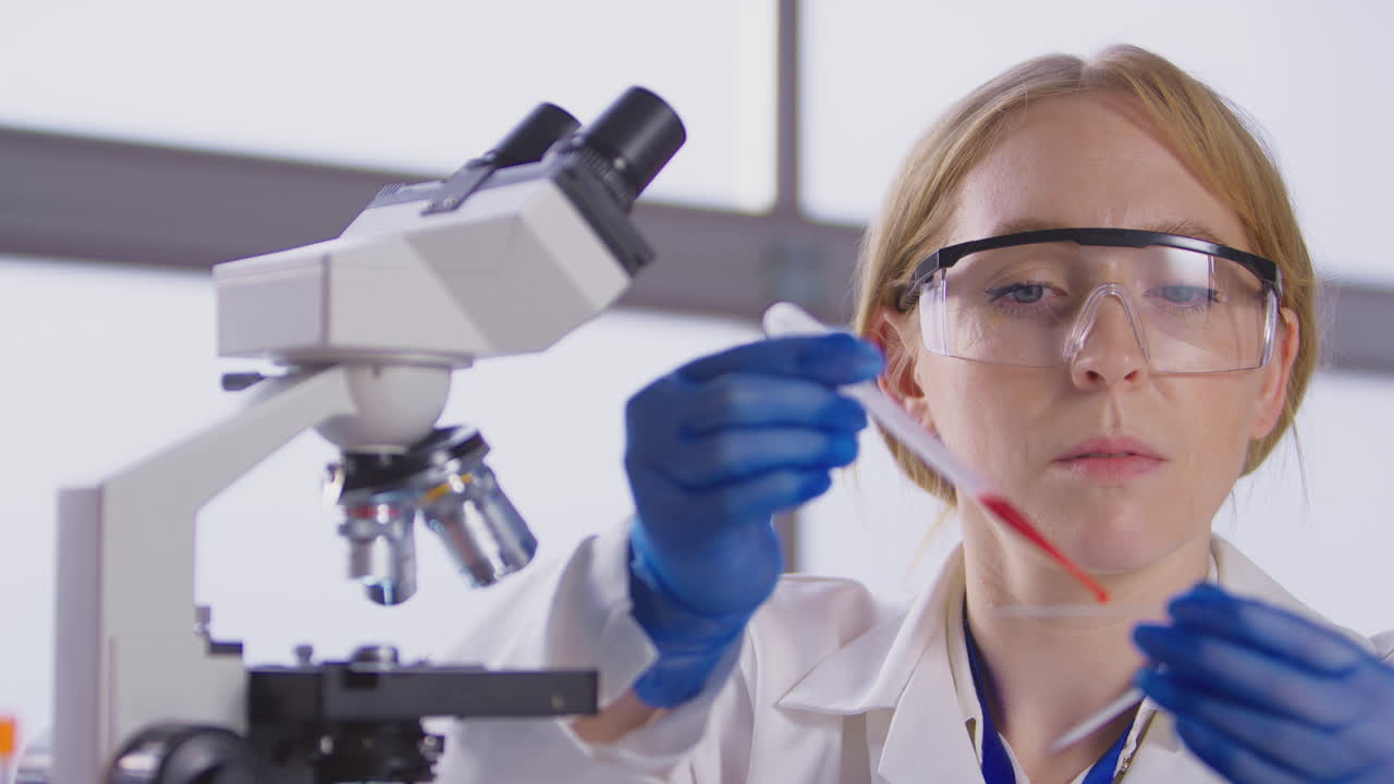 Female Lab Worker Putting Blood Sample Onto Slide For Analysis Under Microscope