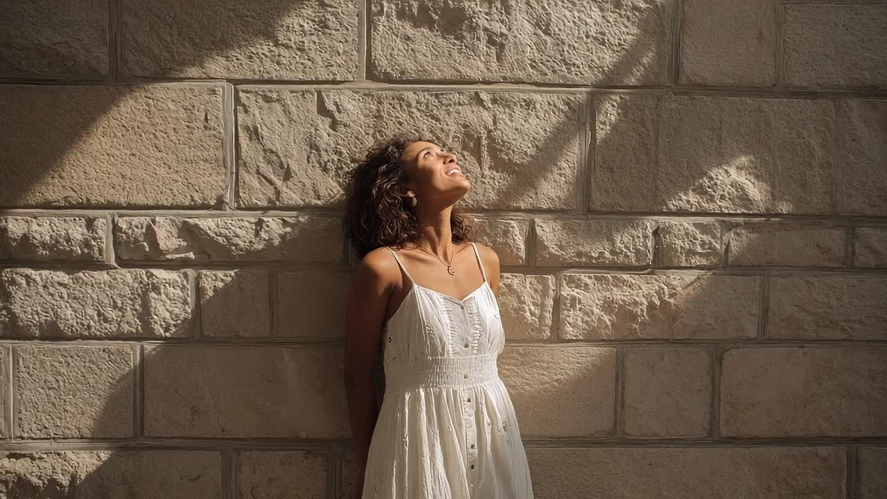 Donna in abito bianco si gode la luce del sole in un contesto urbano vicino a un muro di pietra