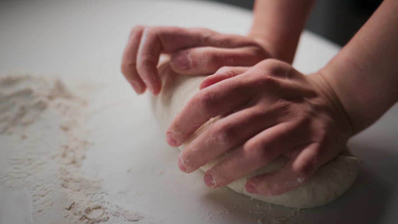 Primo piano di mani che impastano pasta bianca su una superficie infarinata. Le dita premono e piegano l'impasto lentamente. La scena mostra un'attività di panificazione calma e concentrata. Perfetto per ricette, tutorial di cucina o contenuti di panetteria.