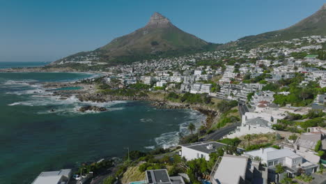 Forwards-fly-above-buildings-in-luxurious-coastal-suburb.-Waves-wasting-rocky-banks.--Pointed-Lions-Head-mountain-against-clear-blue-sky.-Cape-Town,-South-Africa