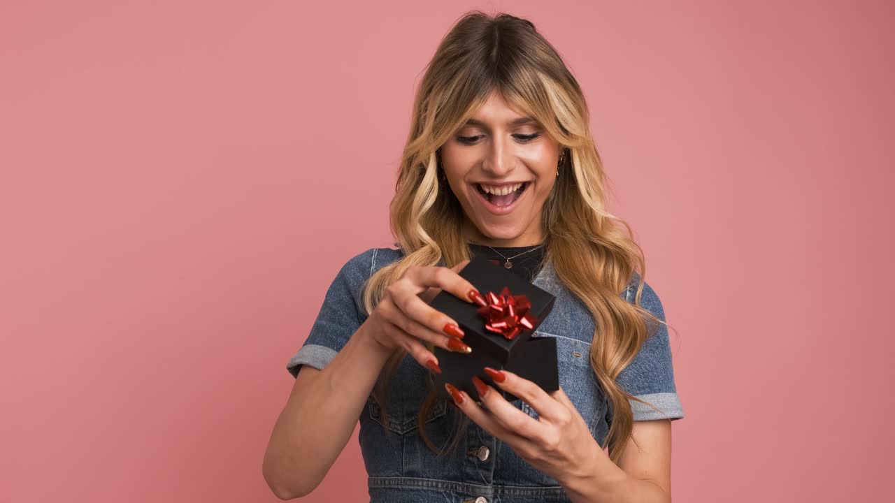Excited transgender woman opening small gift box against pink background