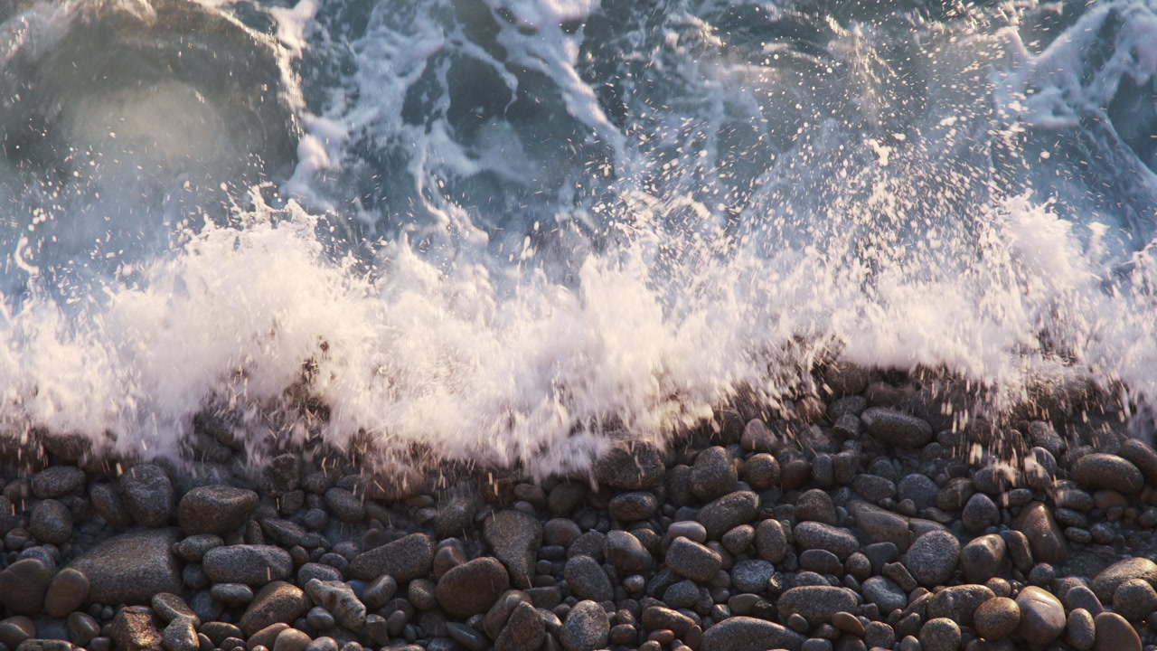Close-up of a wave washing over a shore covered in smooth, sunlit stones and pebbles