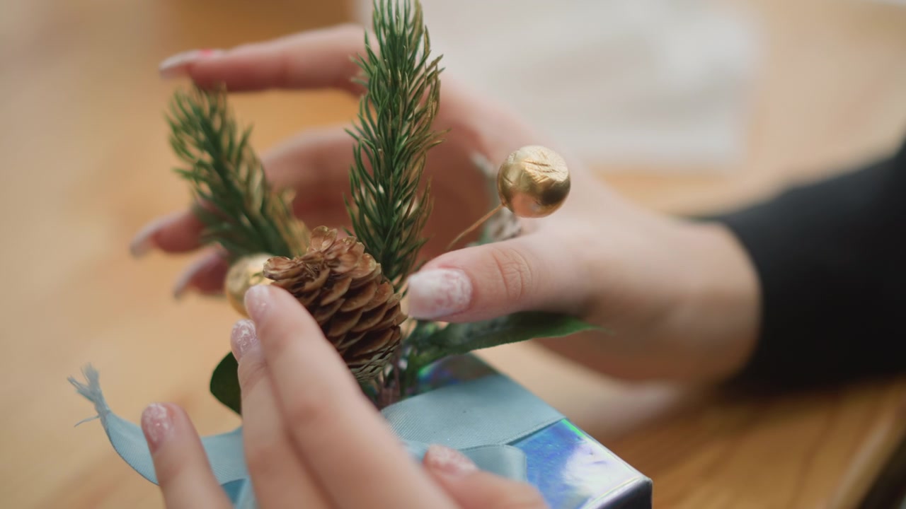 Primo piano che mostra mani che posizionano un rametto sempreverde e una pigna su un regalo di festa incartato con nastro blu, aggiungendo una decorazione naturale e un tocco finale accurato in un ambiente interno caldo.