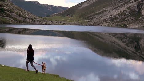 Mujer-Joven-Paseando-Al-Perro-En-Un-Paisaje-Natural-Impresionante-Lago-De-Montañas-Con-Reflejo-Máximo-En-Agua-Quieta