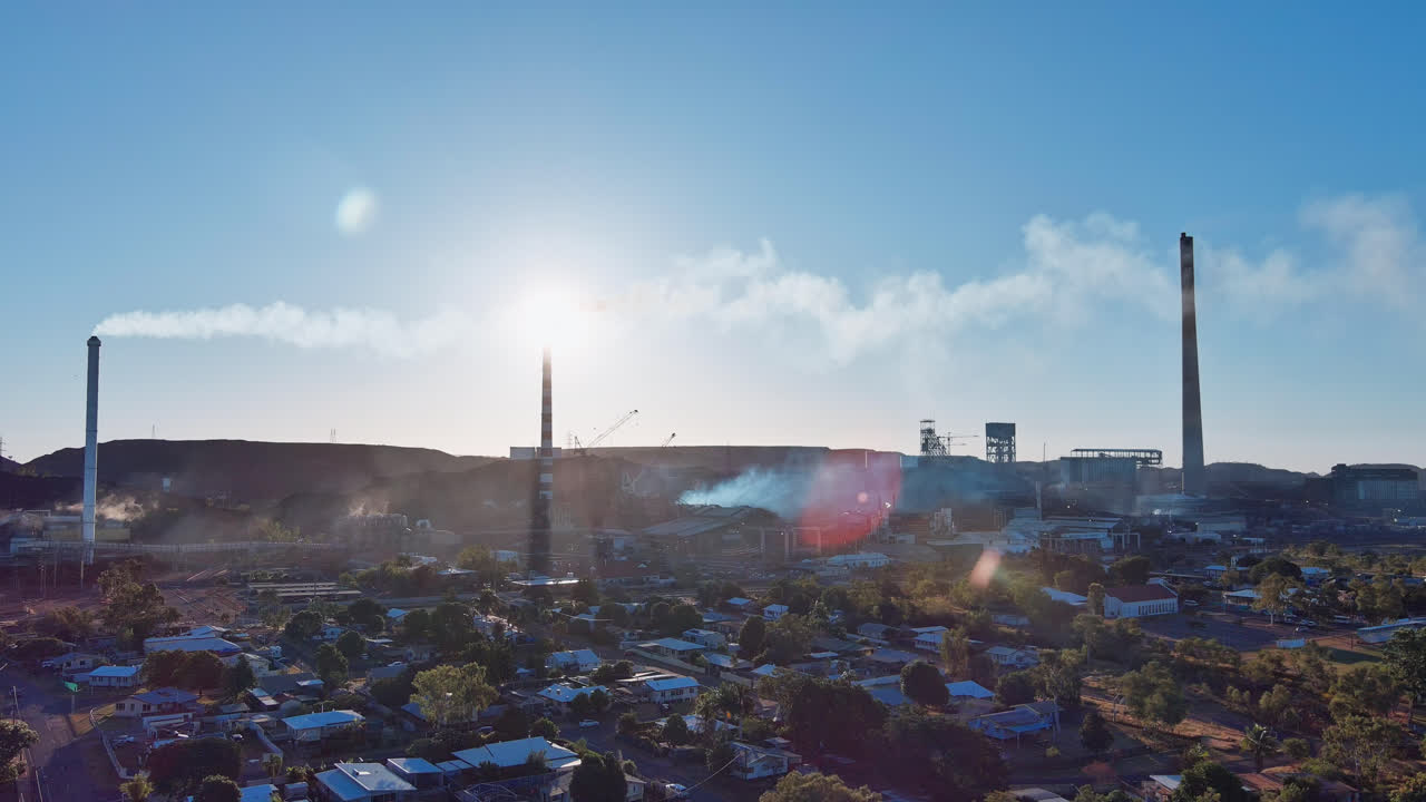 aerial, pista derecha, ciudad de mount isa con las chimeneas de las minas en el fondo con humo en el cielo azul y luces de lente del sol