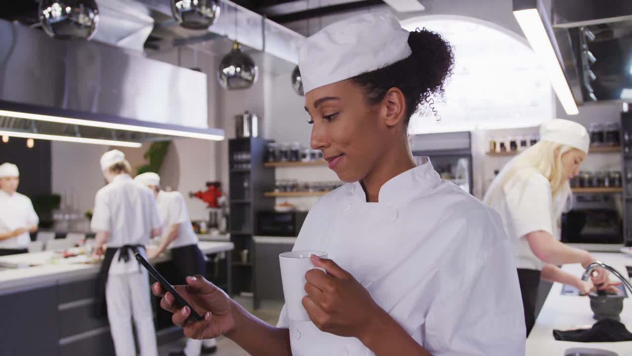 chef afroamericana vestida con ropa blanca en una cocina de un restaurante usando un teléfono y sonriendo
