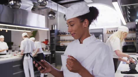 african american female chef wearing chefs whites in a restaurant kitchen using a phone and smiling