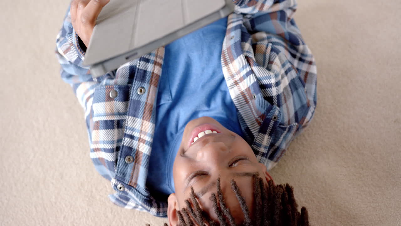 Happy african american boy using tablet lying on floor at home, slow motion