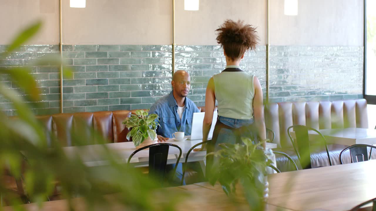 African American server entering in apron checking order, man using laptop reaching for cup at cafe