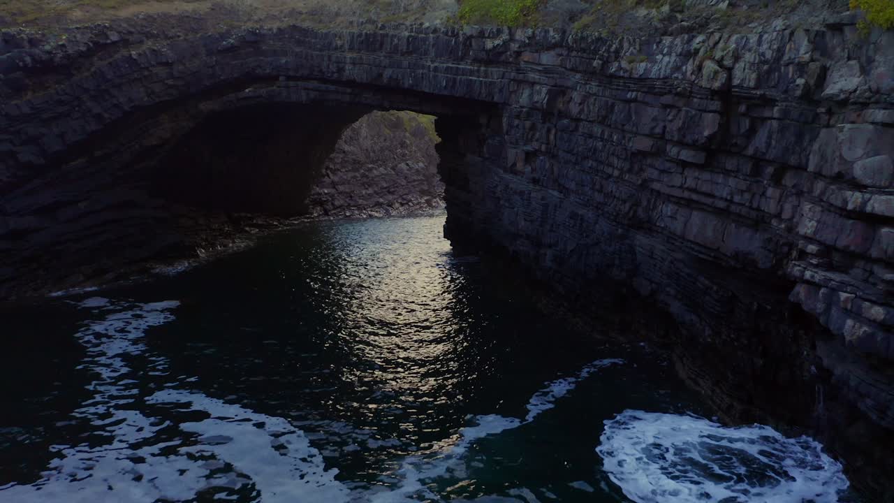 Drone pan follows waves crashing into eroded natural rock bridge arch and flies under towards light glistening, Bridge of Ross Kilkee Ireland