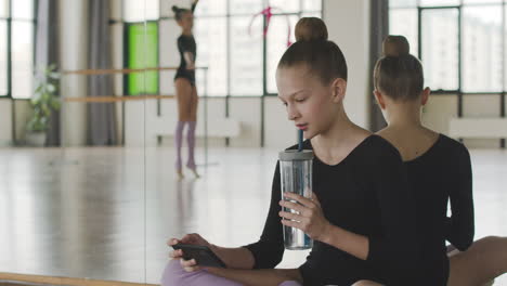 gymnastic blonde girl using smartphone and drinking water sitting on the floor while her friends is dancing holding a band