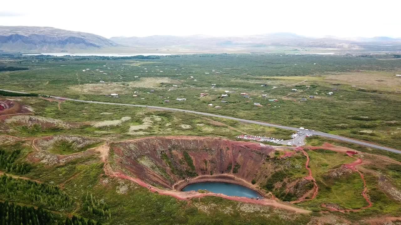 Aerial view capturing Kerid Crater, featuring vibrant red volcanic rock formations surrounding a turquoise lake, with nearby road and parking area, located in the Grímsnes area in south Iceland