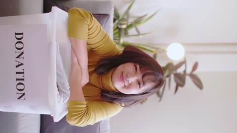 vertical portrait of young asian woman looking at camera holding box with clothes for donation