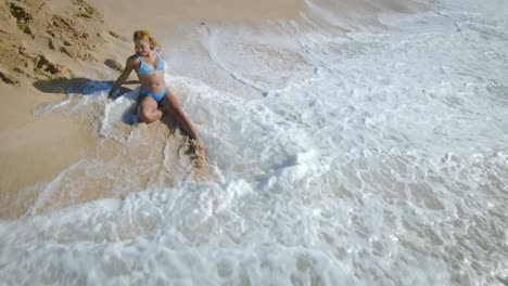 Young-woman-in-blue-bikini-sitting-on-ocean-beach-in-tropical-Paradise-Island-with-turquoise-water-waves,-white-sand-in-sunny-weather