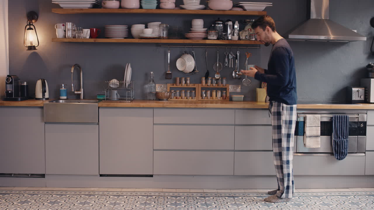 Giovane uomo felice che balla in cucina al mattino, vestito, con smartphone e caffè.