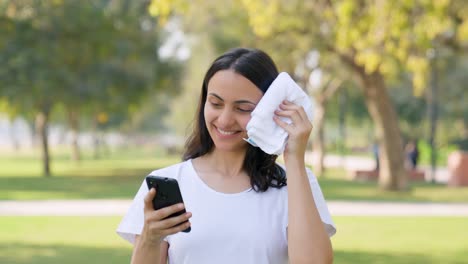 Mujer-India-Desplazando-El-Teléfono-Mientras-Se-Limpia-El-Sudor-En-Un-Parque-Por-La-Mañana