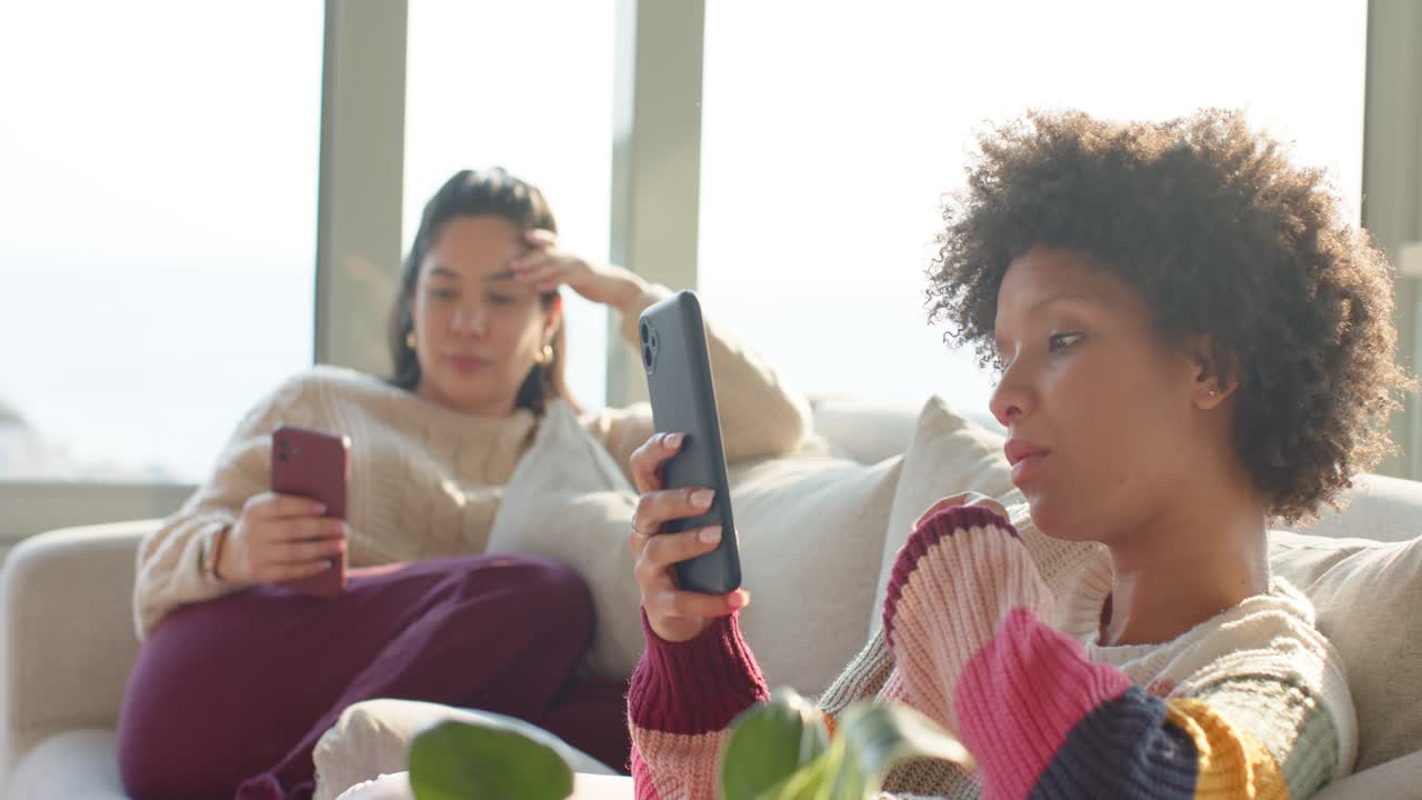 Diverse female friends lifting smartphones and scrolling through messages on light sofa by window
