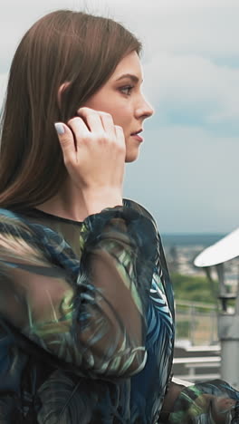 tranquil young woman with long hair in cute dress with colorful print enjoys large city view leaning on footbridge railing slow motion close view