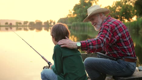 rear view of a teen boy sitting with his grandfather on the lake pier, talking and fishing together