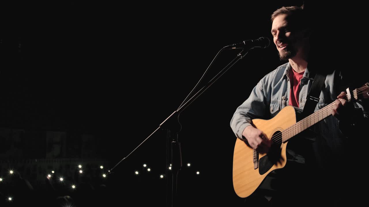 un músico está tocando la guitarra en un gran escenario con miembros de la audiencia sosteniendo linternas en el fondo. el auditorio está lleno de hermosas luces parpadeantes.