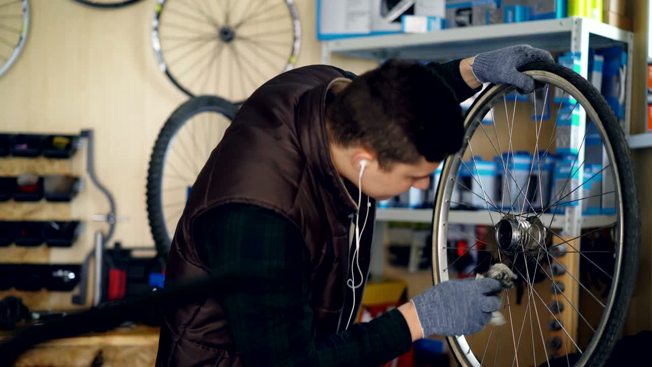 el joven reparador está limpiando la rueda de la bicicleta dentro y fuera de una pieza de tela sucia y escuchando música con auriculares. profesión y concepto de la gente.