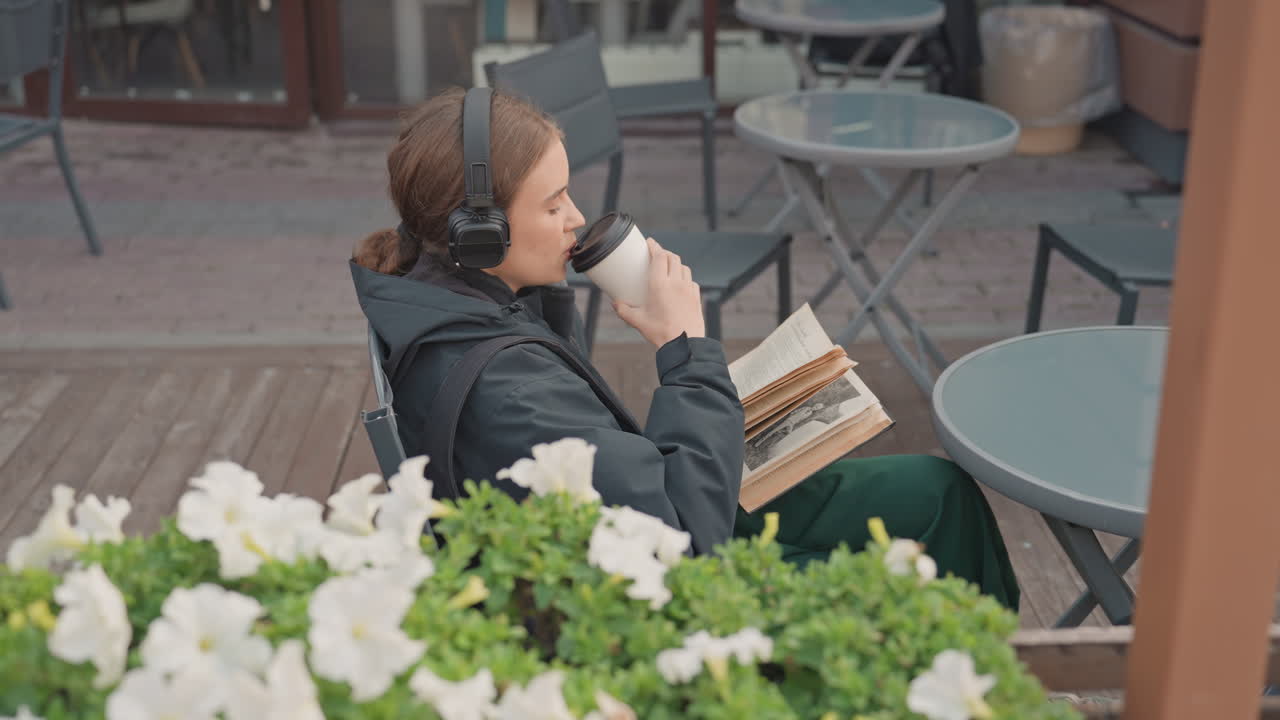 Kvinde, der læser på en udendørs café med hovedtelefoner på, iført en mørk jakke, mens hun drikker kaffe fra en to-go kop og bladrer i en paperback-bog på et træterrassebord omgivet af hvide blomster og potteplanter