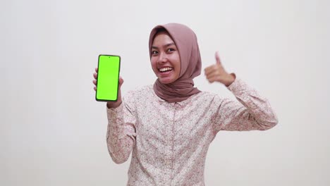 happy young asian muslim girl showing pointing presenting a green screen of cell phone display, suggested something on her phone