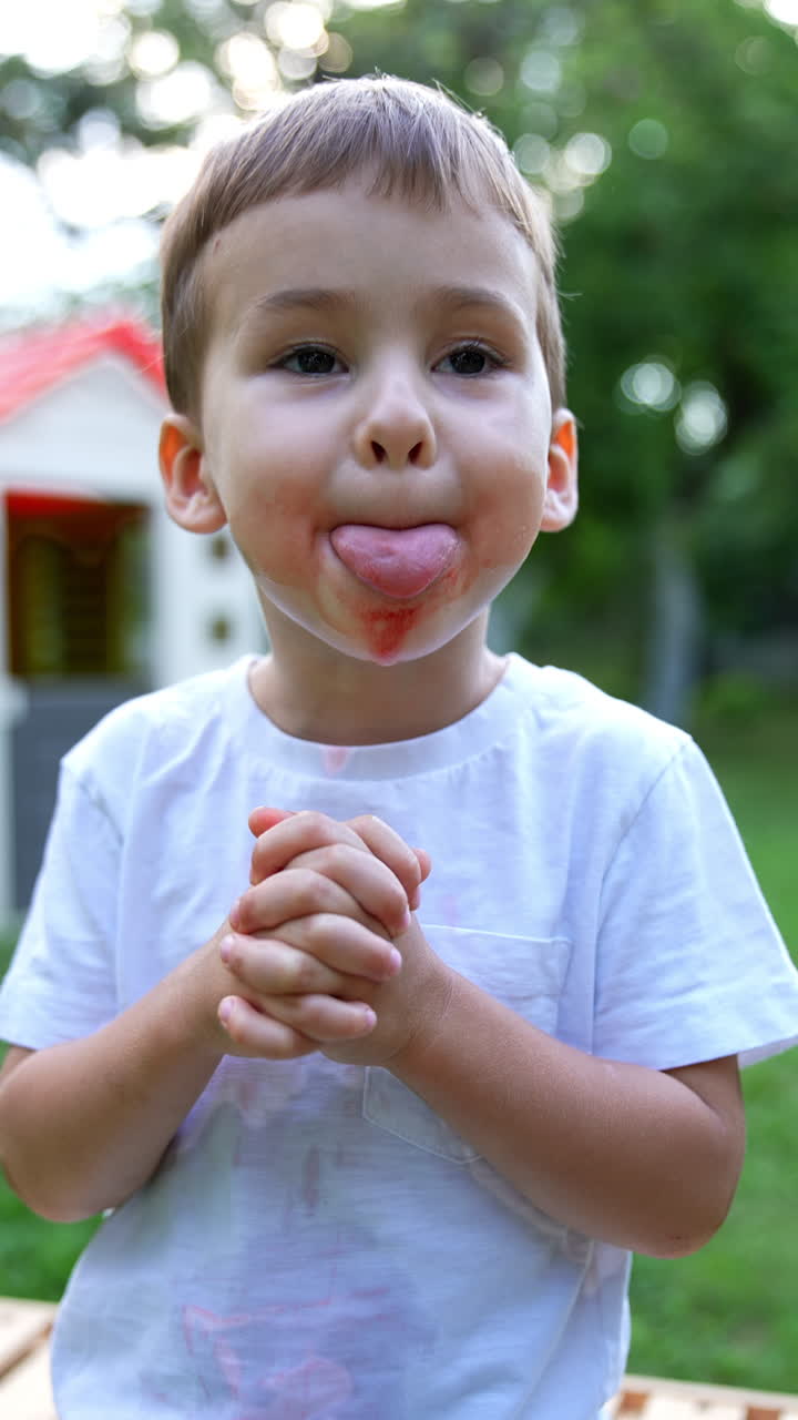 Little kid sitting outdoors with a smudged face. Cute child has eaten a watermelon and has dirty face and hands. Vertical video.
