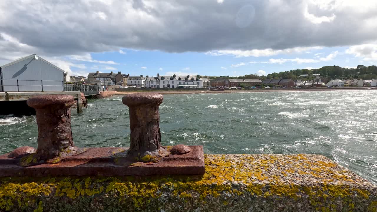 Rusty mooring bollards on a weathered pier overlook choppy harbor waters, with coastal village houses in the background under dynamic, partly cloudy daylight