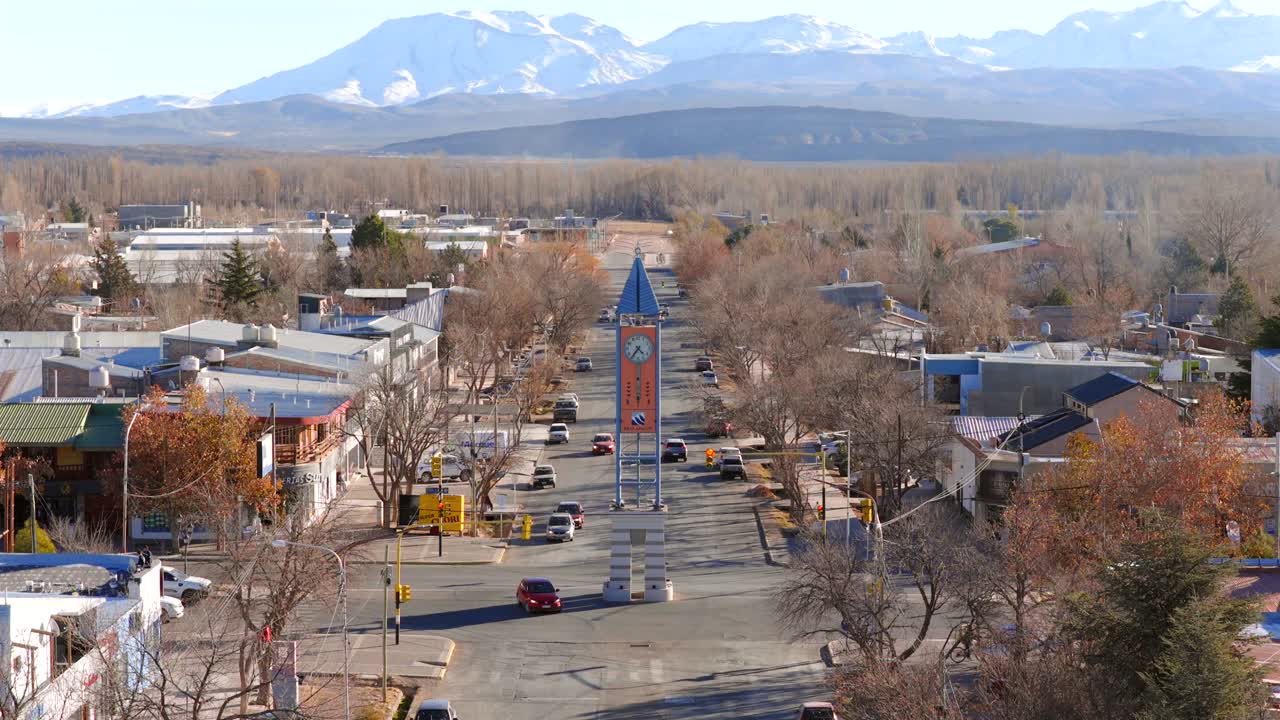 Drone slowly descends over Malargüe, Mendoza, Argentina, featuring the iconic clock tower monument in the town center, framed by a straight avenue, leafless trees, and the towering Andes behind.