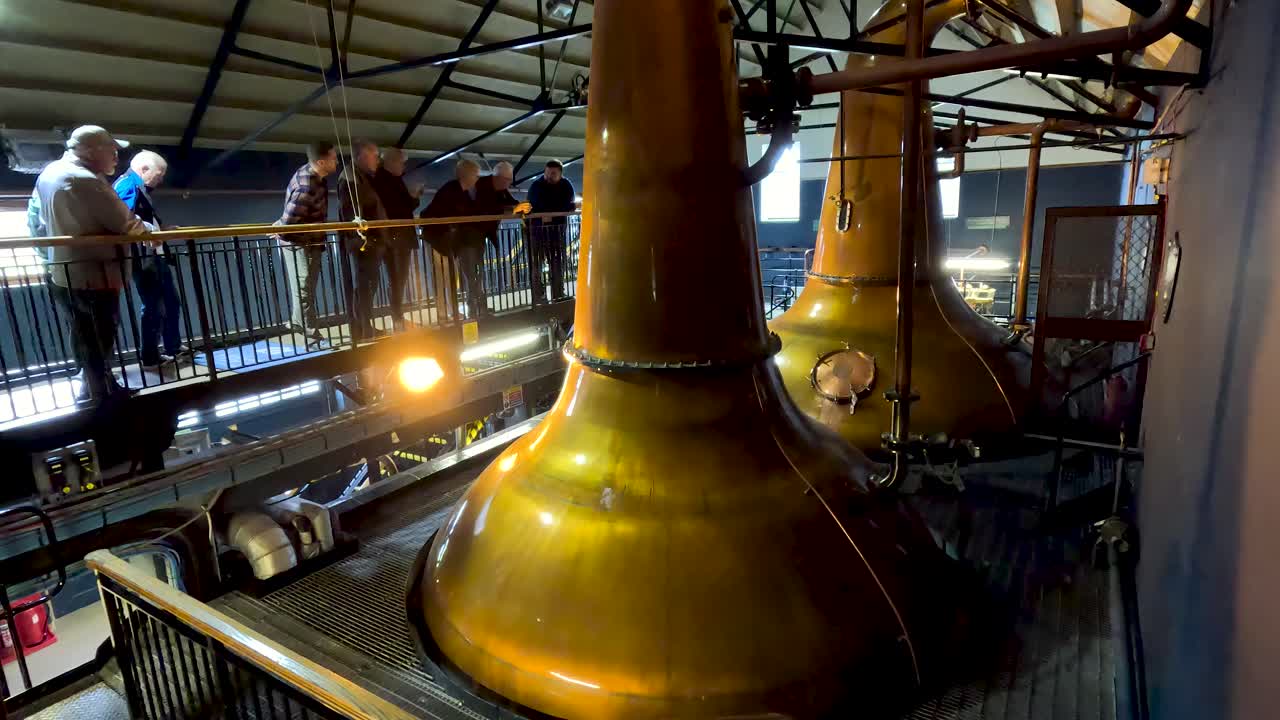 Tour group observes copper pot stills inside industrial distillery, warm lighting, static wide shot