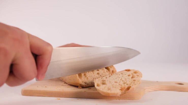 Close-up shot of hands using a large knife to slice sourdough bread on wooden board.