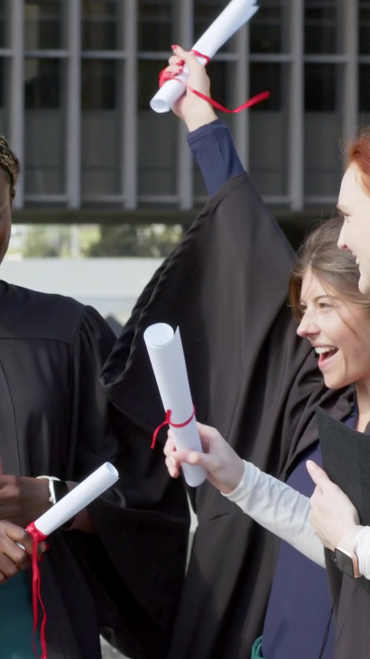 Vertical video: Raising diplomas after ceremony, three grads in gowns celebrating at campus plaza