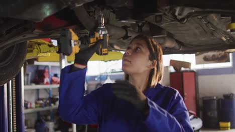 Mecánica-Femenina-Usando-Un-Taladro-Eléctrico-Y-Trabajando-Debajo-De-Un-Automóvil-En-Una-Estación-De-Servicio-De-Automóviles