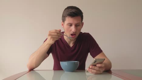 mann reagiert auf inhalte auf dem mobiltelefon, während er müsli isst, sitzt an einem glastisch in minimalistischer inneneinrichtung, immer noch mittlere aufnahme auf augenhöhe mit blick auf die kamera