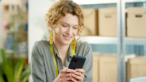 Close-up-view-of-caucasian-designer-woman-sitting-at-desk-texting-on-smartphone-in-clothing-shop-warehouse