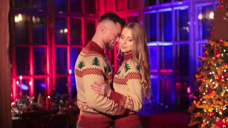 hugging couple in similar knitted warm sweaters stand in the country house. pair in love spending christmas holidays outside the city.
