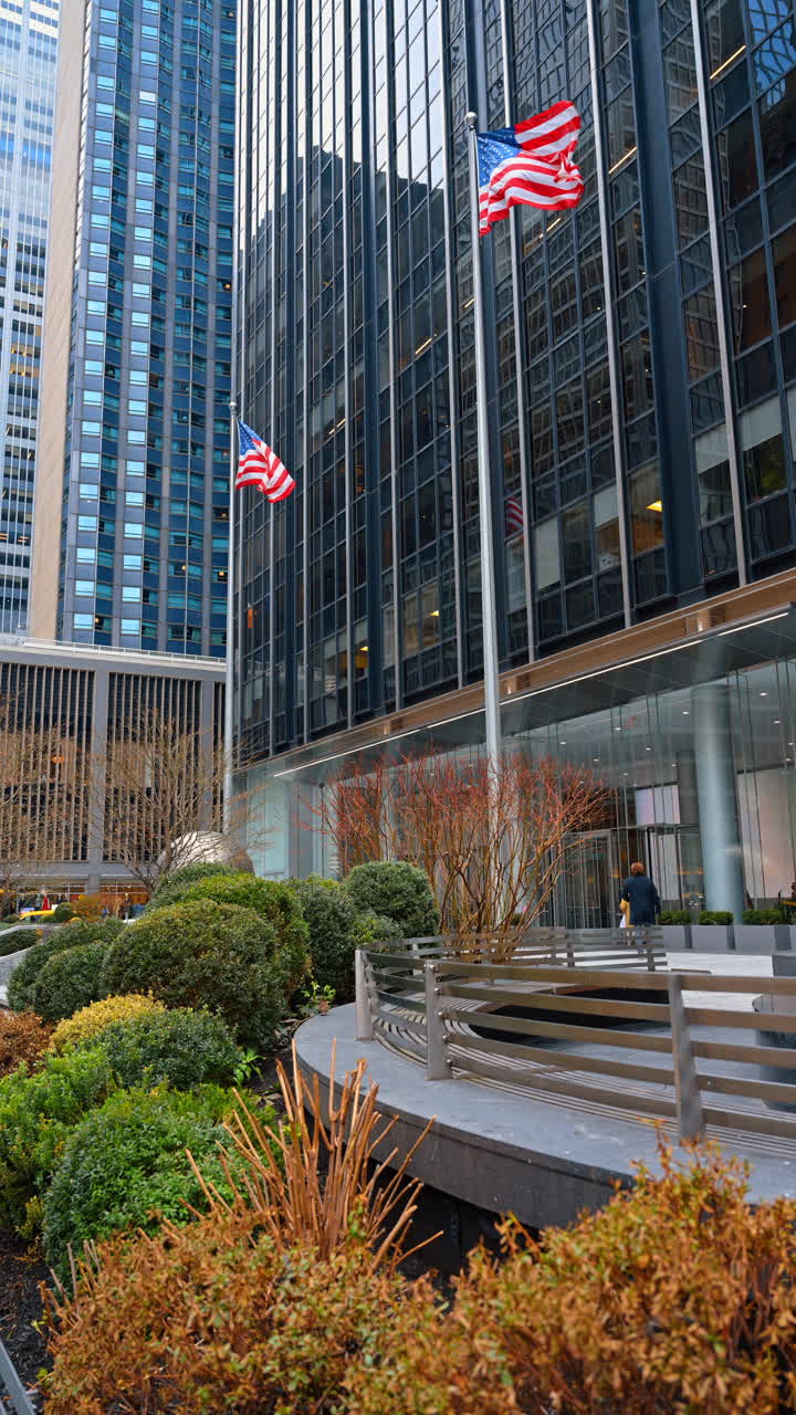 Bandiere americane di fronte alla piazza di un moderno edificio per uffici di Manhattan. Bandiere degli Stati Uniti su aste all'esterno di un grattacielo di vetro con cespugli da giardino e persone a New York City.