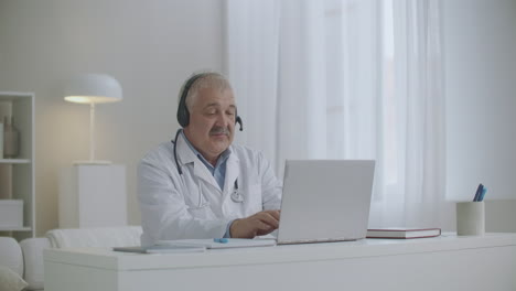 médico de cabelos grisalhos está conversando on-line pela internet usando laptop em seu escritório se comunicando com colegas e pacientes