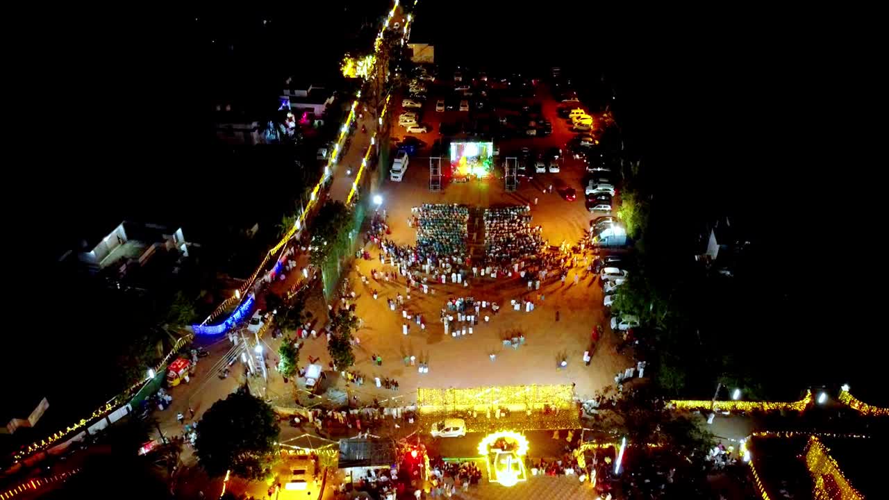 En levende droneoptagelse fra luften af en overfyldt udendørs natfestival eller koncert i Indien, med en stærkt oplyst scene, farverige lysshow og store grupper af mennesker i traditionel påklædning