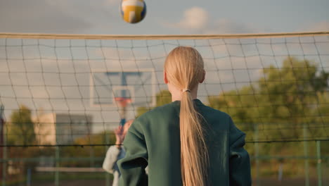 vista trasera de un jugador de voleibol con capucha verde participando en un juego cerca de la red con cabello largo y rubio, cancha deportiva al aire libre y edificio en el fondo bajo un cielo despejado