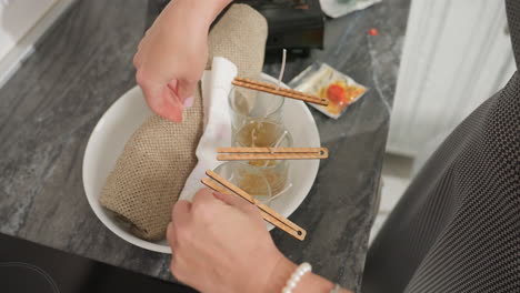 hand view of woman inserting tissue paper between three glass cups and sack bag inside white bowl to stabilize candle containers, focusing on precise setup for candle crafting on kitchen countertop