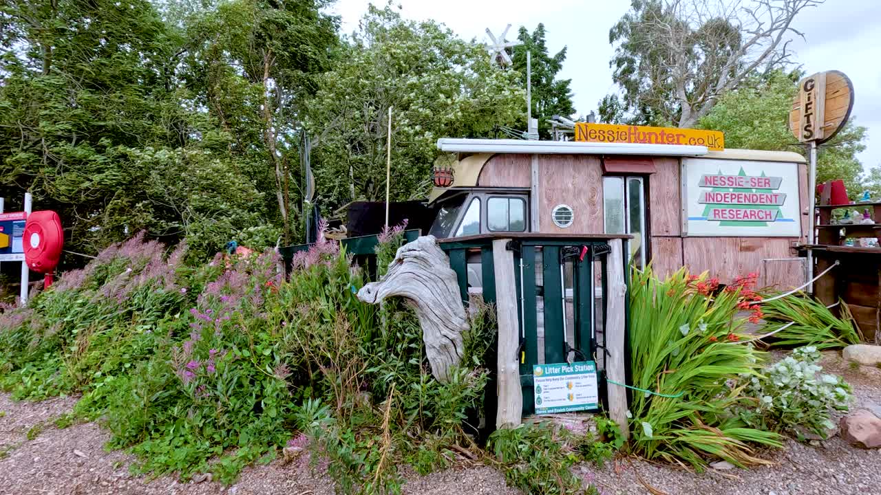 A person walks along a pebbled lakeshore, passing a quirky tourist shop surrounded by greenery on Dores Beach, under overcast daylight in the Scottish Highlands