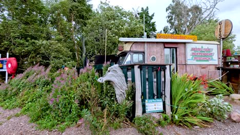 a person walks along a pebbled lakeshore, passing a quirky tourist shop surrounded by greenery on dores beach, under overcast daylight in the scottish highlands